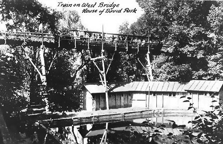 With all the colonists had to do to open the Eden Springs Park in 1908 they found time to construct a high railroad trestle over the creek valley in the center of the park.  This trestle was at the west end of the valley and is shown in the earliest pictures of the park.  It is not clear today how this trestle was used before the east trestle was built a short time later.  Trains may have run forward over the trestle in one direction and in reverse over it in the other direction.  Another possibility is that the train looped on each side of the trestle.  The west trestle ran just to the west of a pond made by impounding the creek that ran through the Eden Springs Valley.  In the foreground are buildings that were used as changing rooms for swimming.  The train in this image is heading south from the north depot to the south depot.  This was most common direction of travel during the history of the railroad.  In later years park visitors could see trains pass on this trestle behind the beer garden stage while watching shows at the park.  The Eden Springs creek ran from the pond, over a spillway, under this trestle and out of the park.